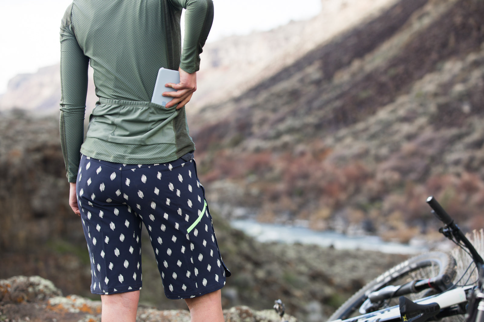 A person wearing Wild Rye shorts and a long-sleeve shirt, placing a phone in their back pocket, with a scenic outdoor background.