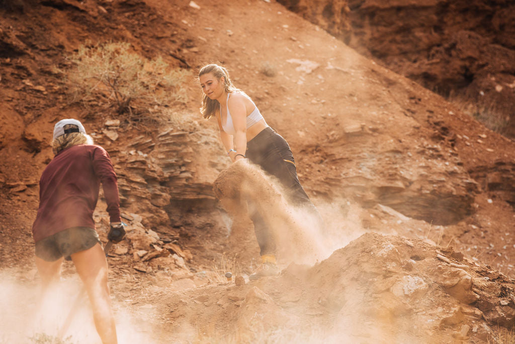 Two women working on a dirt mound in a dusty landscape during a mountain biking event.