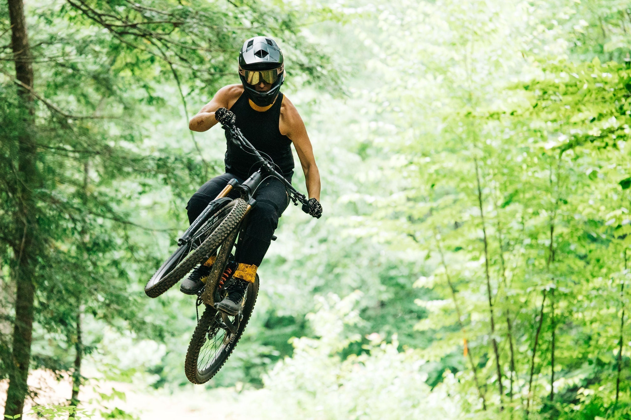 A person in a black tank top and helmet jumping on a mountain bike in a forest.