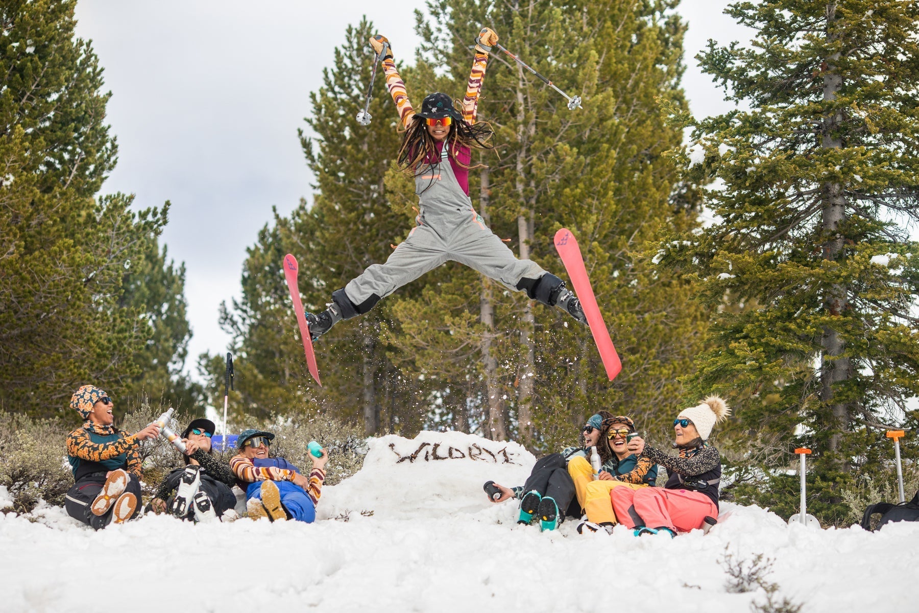 A skier jumping in the air with a group of people sitting in the snow, surrounded by trees.