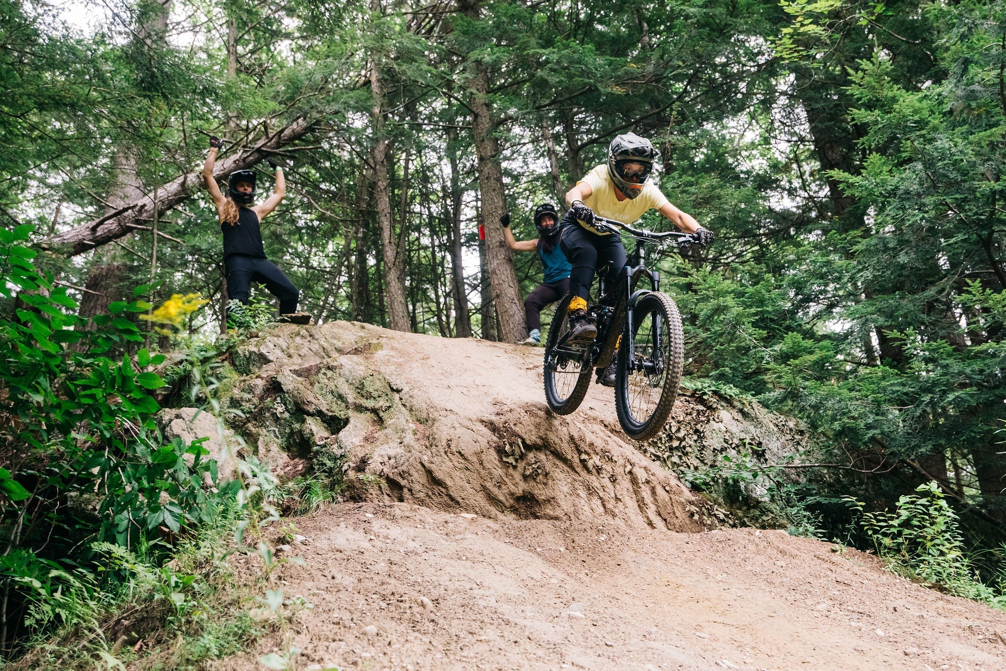 A mountain biker jumping off a dirt ramp with two spectators cheering in the background.