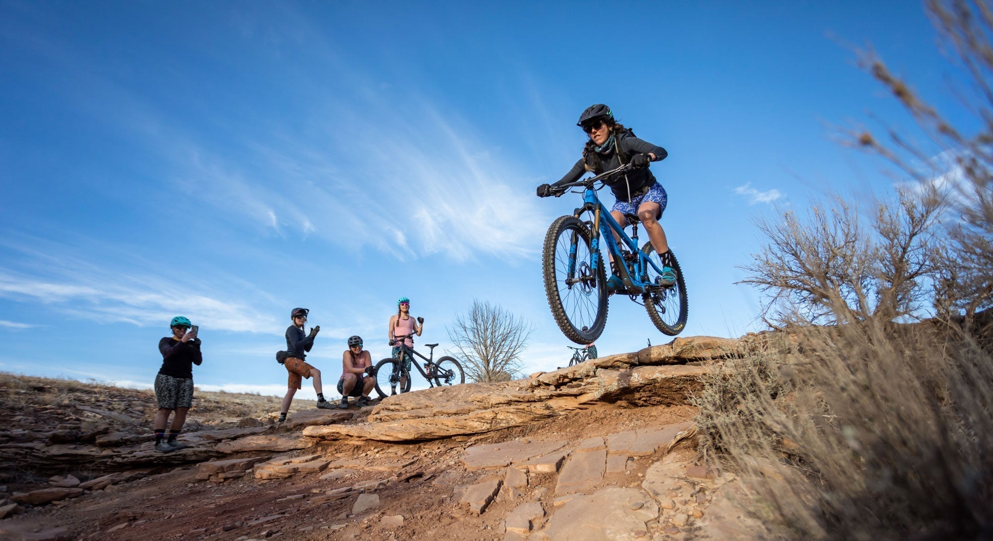 A woman jumping on a mountain bike while others watch in the backcountry