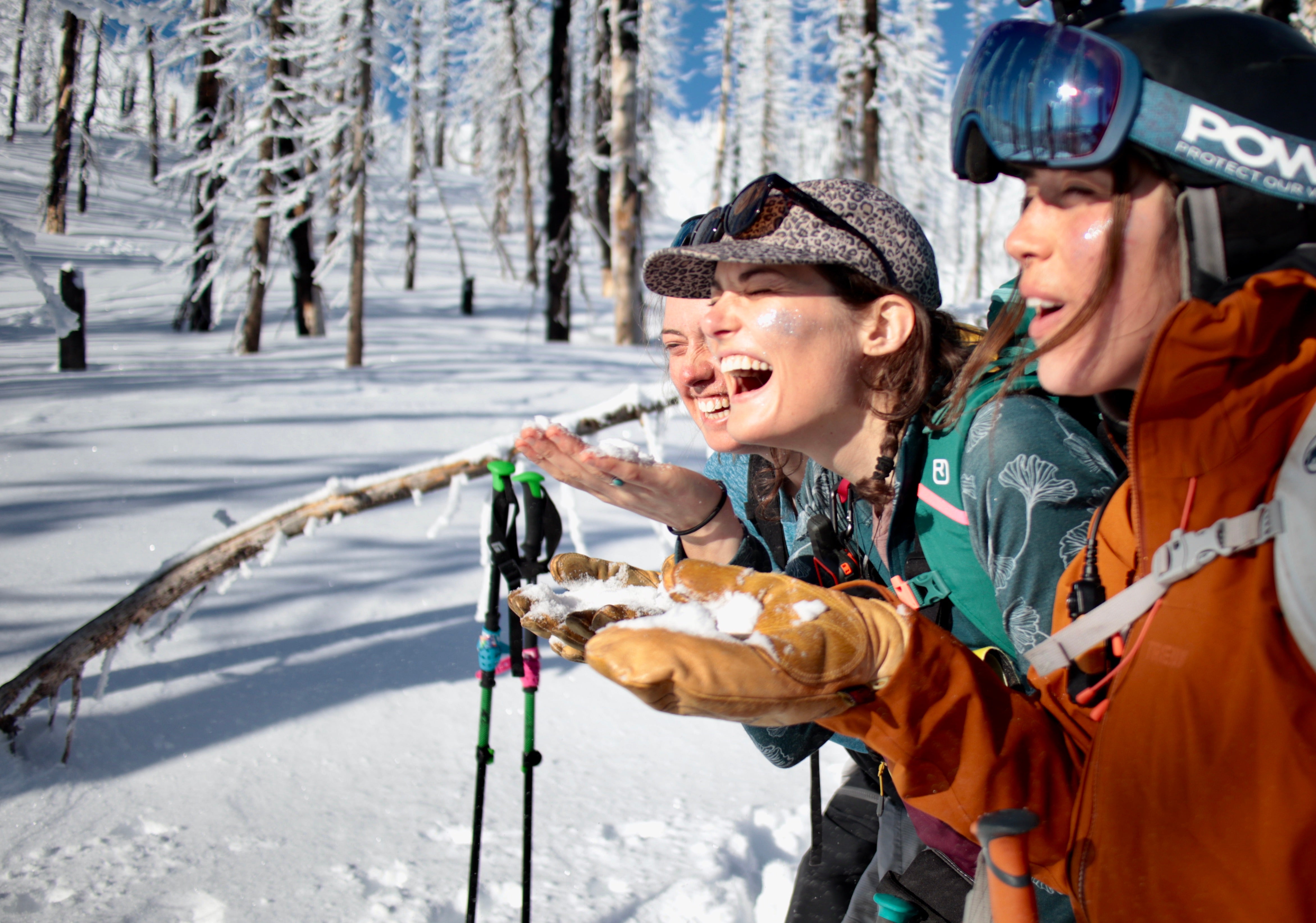 Three women smiling and playing in the snow, surrounded by snowy trees.