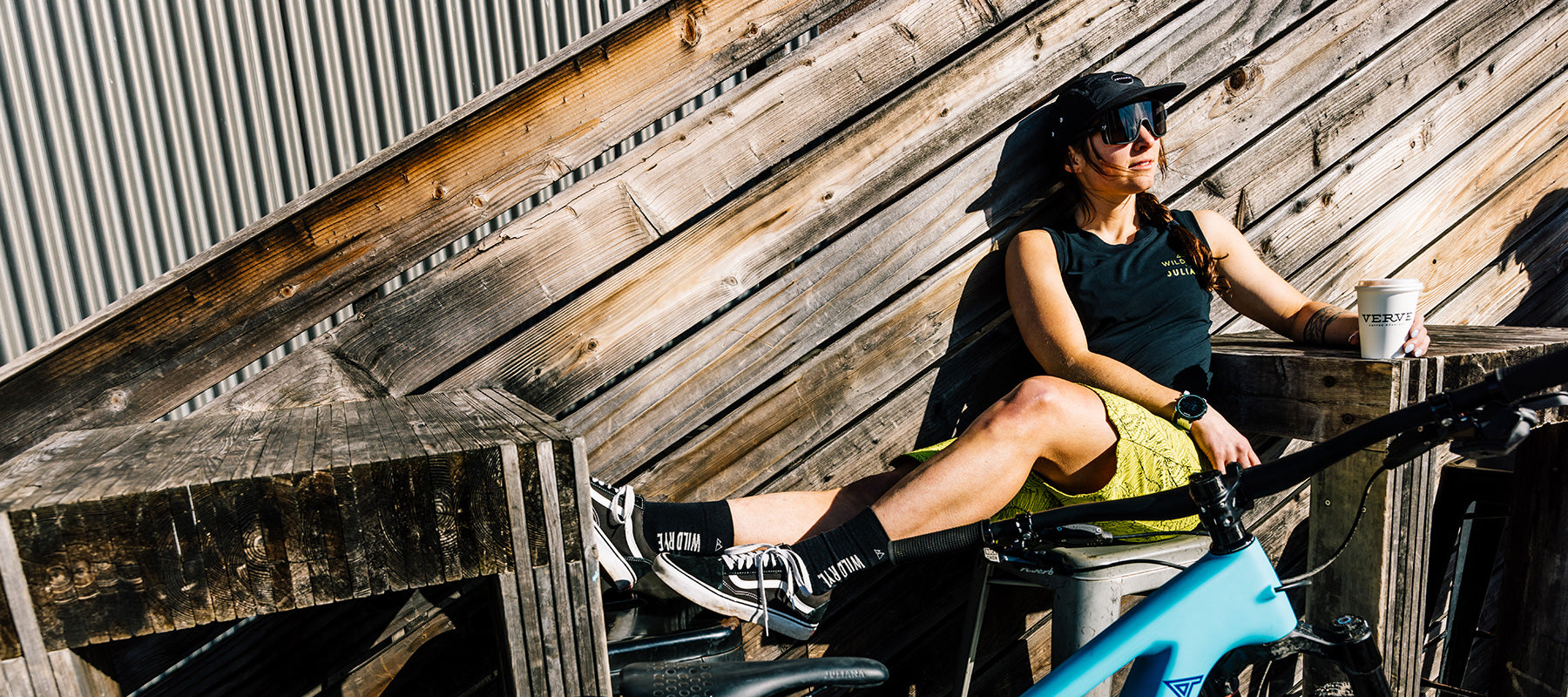 A woman in a tank top and shorts sitting on a wooden structure with a bike, holding a coffee cup.