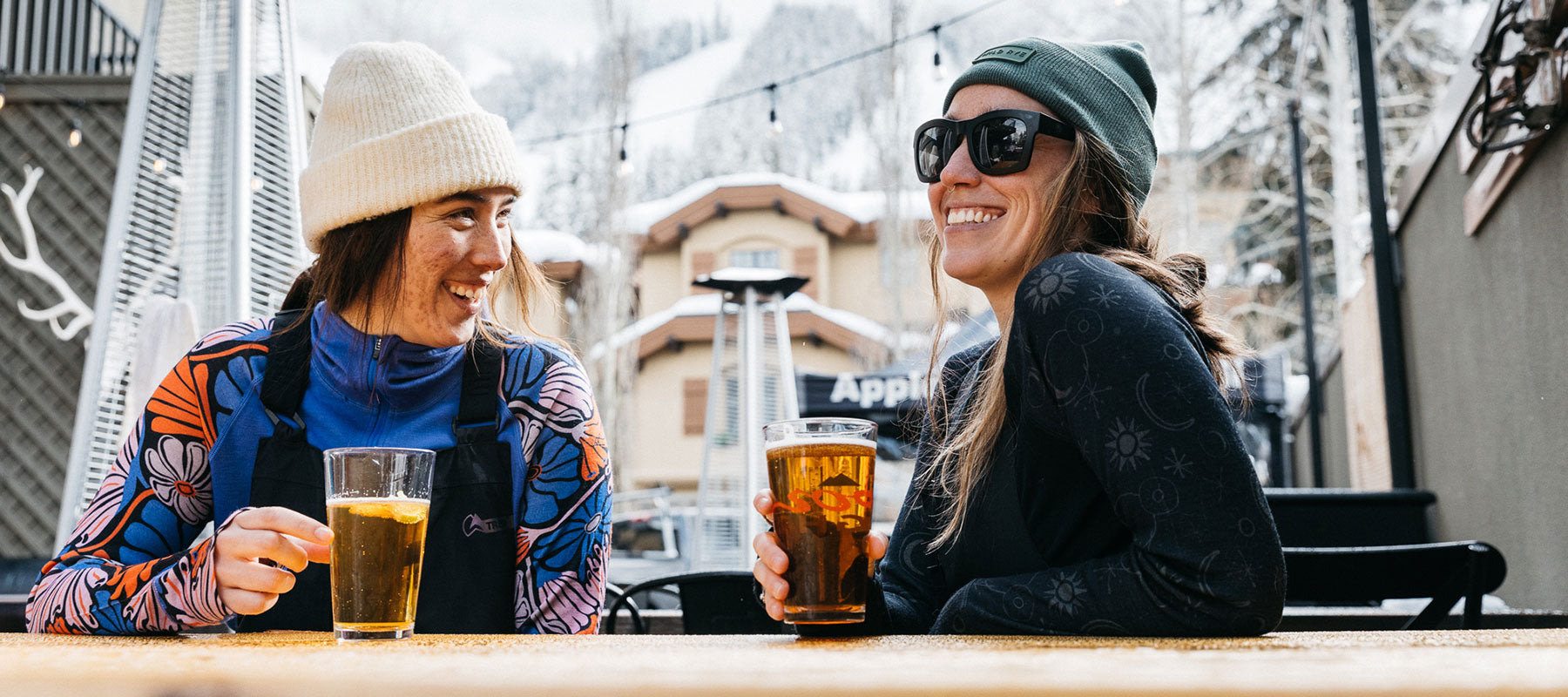 Two women wearing colorful 100% merino baselayers and winter hats, sitting outdoors at a table with drinks, smiling and talking, with snowy mountains in the background.