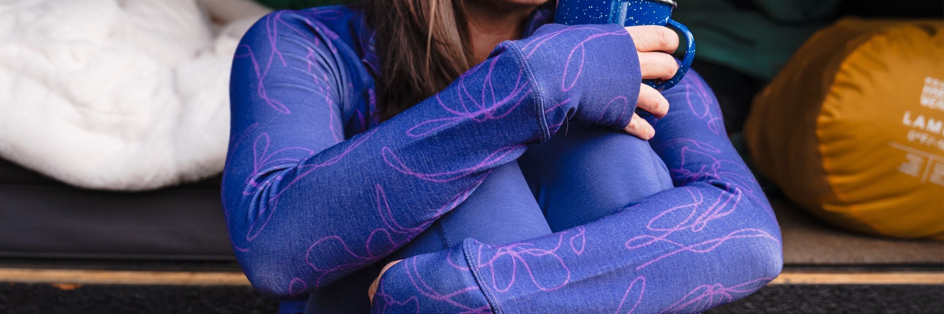 Three women wearing colorful baselayers and holding mugs, standing indoors by a large window with a snowy outdoor scene visible.
