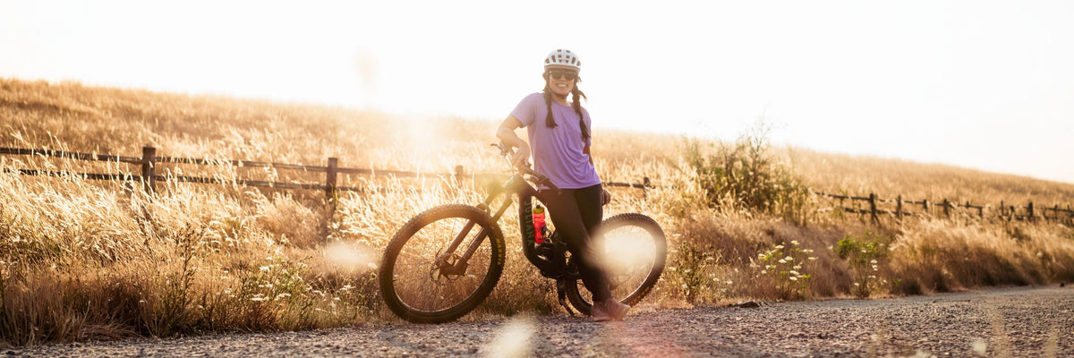 Three women carrying mountain bikes uphill on rocky terrain, wearing cycling bottoms and helmets.