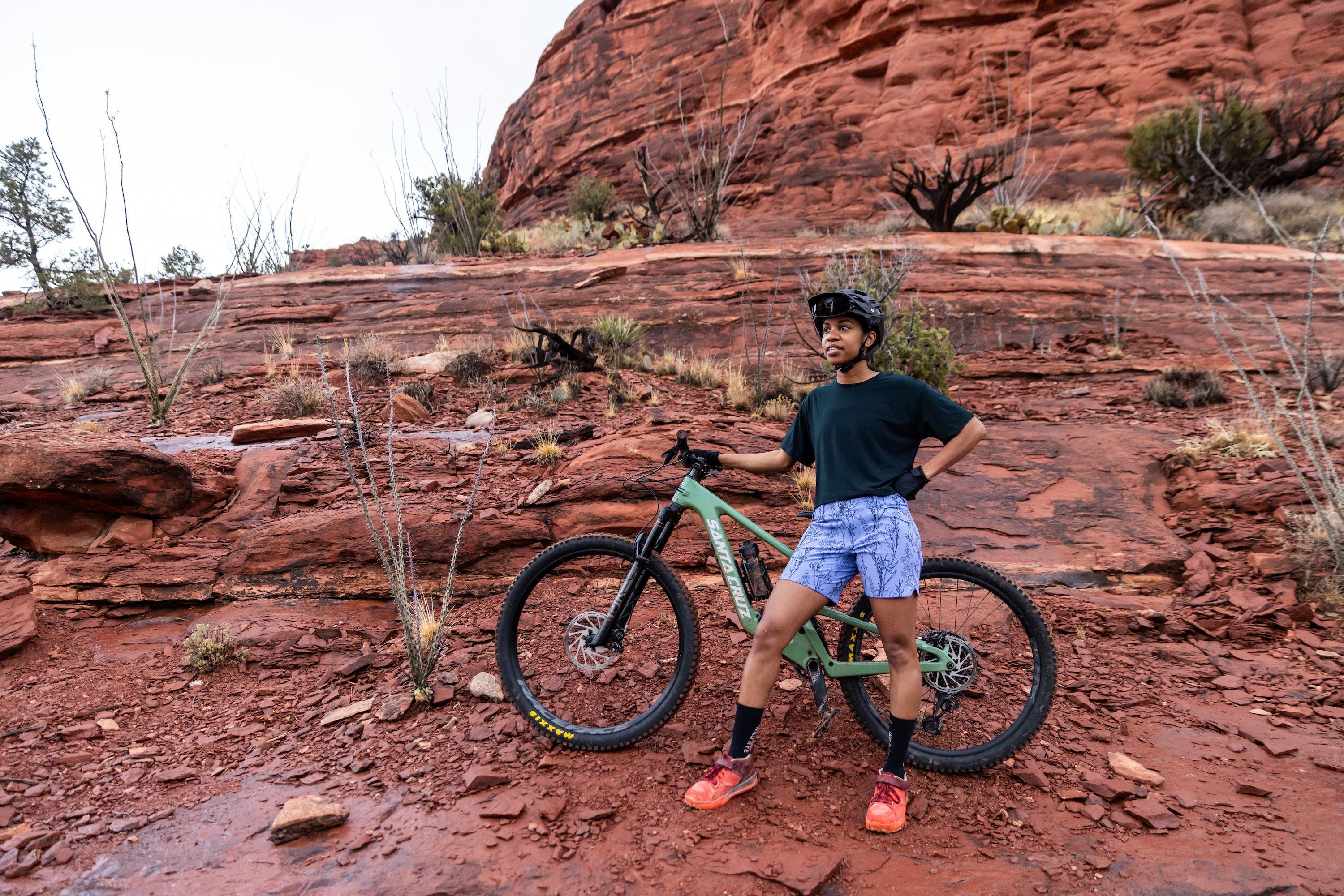 Person wearing a helmet and gloves standing with a green mountain bike on red rocky terrain with sparse vegetation and red rock formations in the background