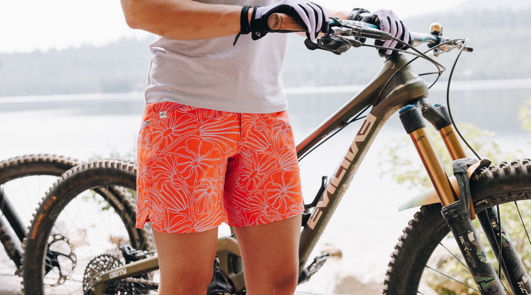 Person wearing orange floral shorts and white gloves holding the handlebars of a mountain bike with 'EVIL' branding on the frame, outdoors near a lake.