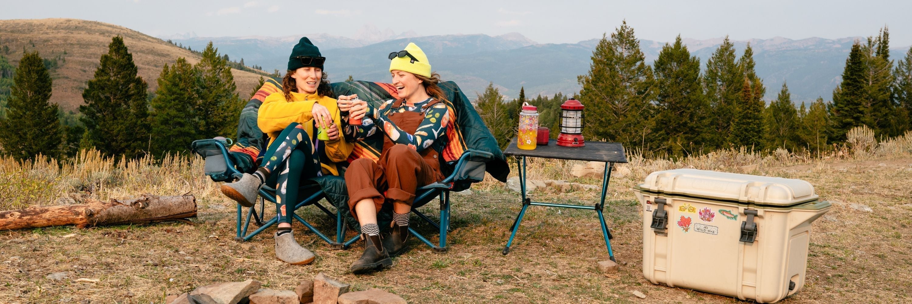 Two people outdoors in front of garages; one rides a white bicycle wearing an orange cap and gray overalls, the other crouches on a skateboard holding the bike's rear rack, both smiling.