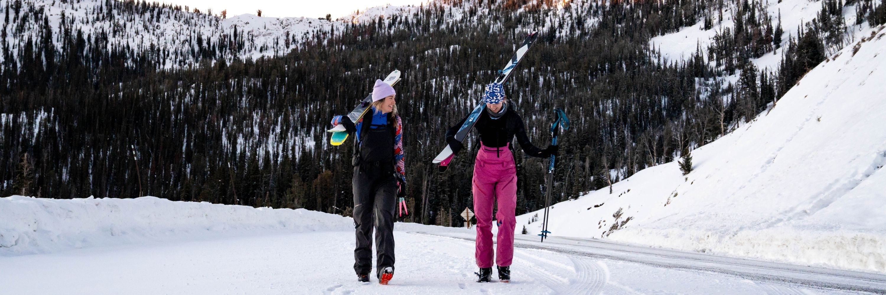 Two women carrying skis and ski poles walking on a snowy mountain road with forested hills in the background