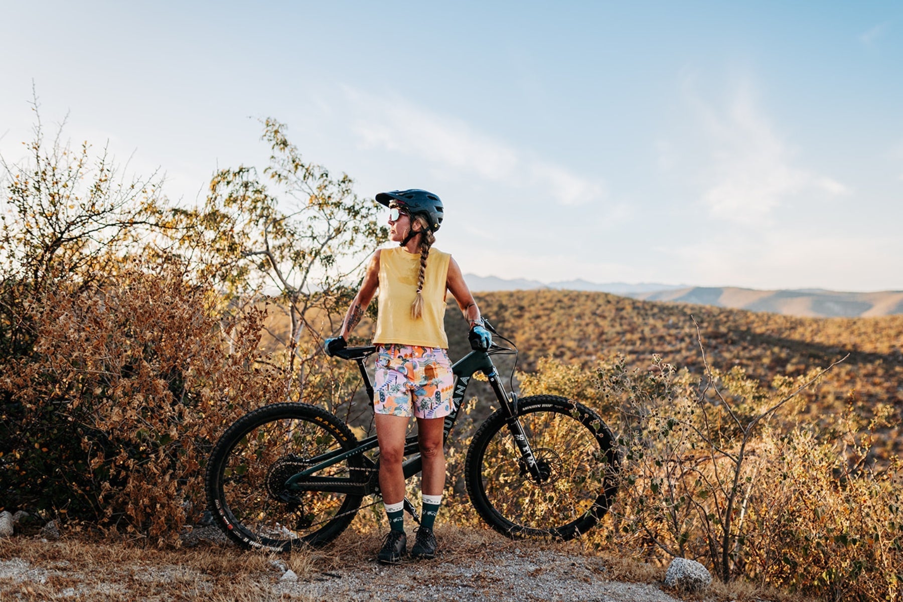 Woman wearing a helmet and colorful shorts standing with a mountain bike in a dry, hilly landscape under a clear sky.