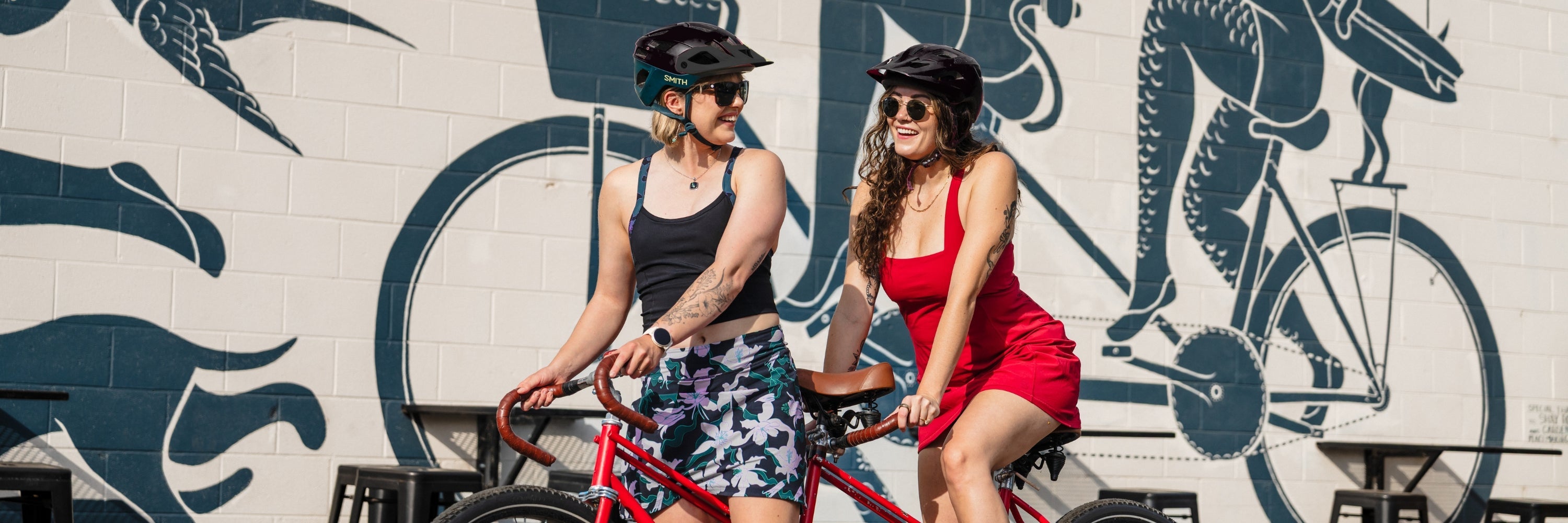 Two women sitting on a sailboat by the ocean, one wearing a teal and black patterned dress, the other in an orange and white dress, enjoying a sunny day.