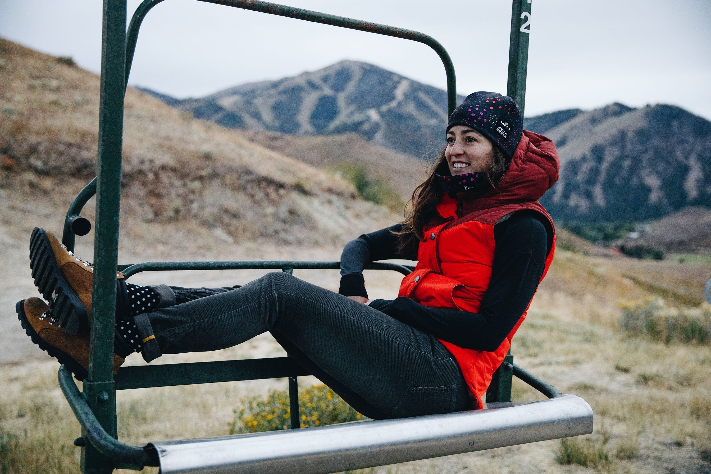 Woman wearing a red vest, black long sleeve shirt, black beanie with pink dots, and brown boots sitting on a ski lift chair with mountains in the background.