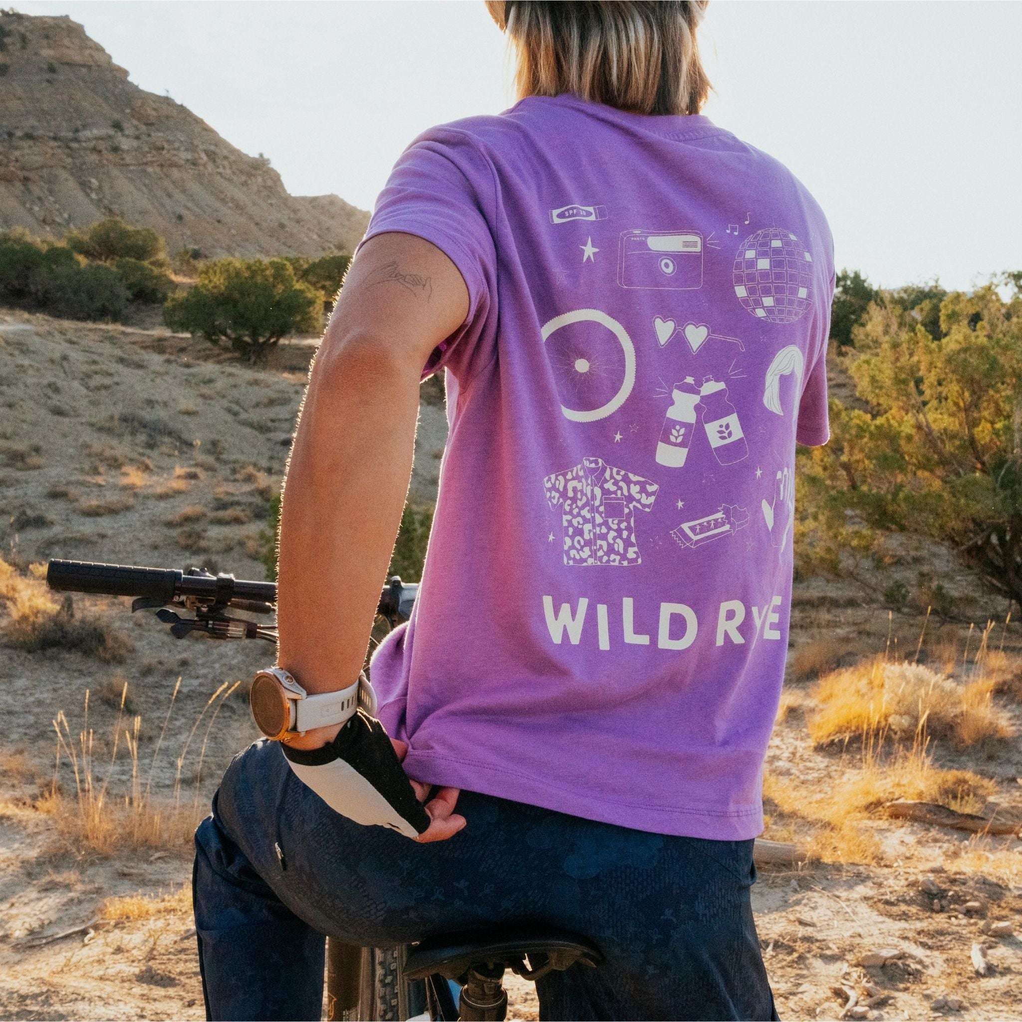 Person on bike, showing the back of a purple shirt with fun icons of wigs, sunglasses, cameras, and disco ball