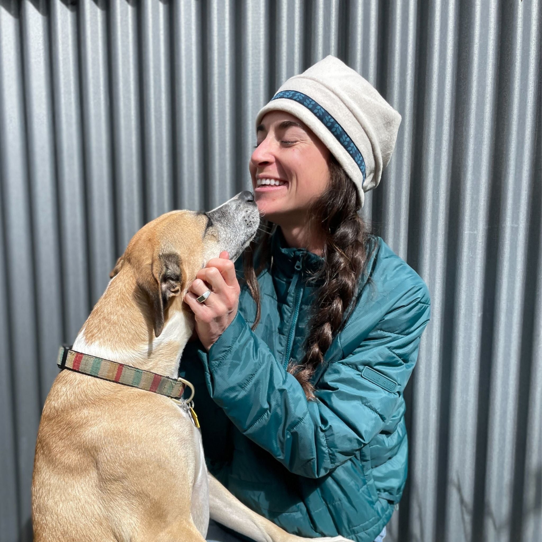 Woman in a green jacket and white beanie interacting with a dog against a corrugated metal background
