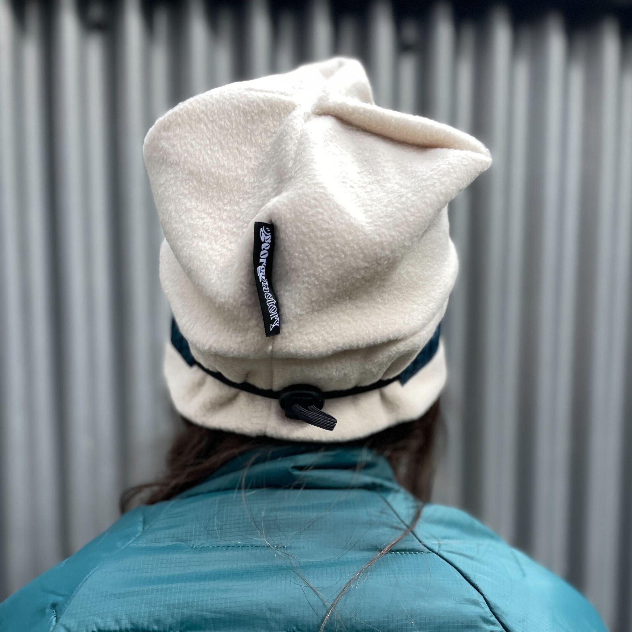 Person wearing a white beanie with a logo against a corrugated metal background