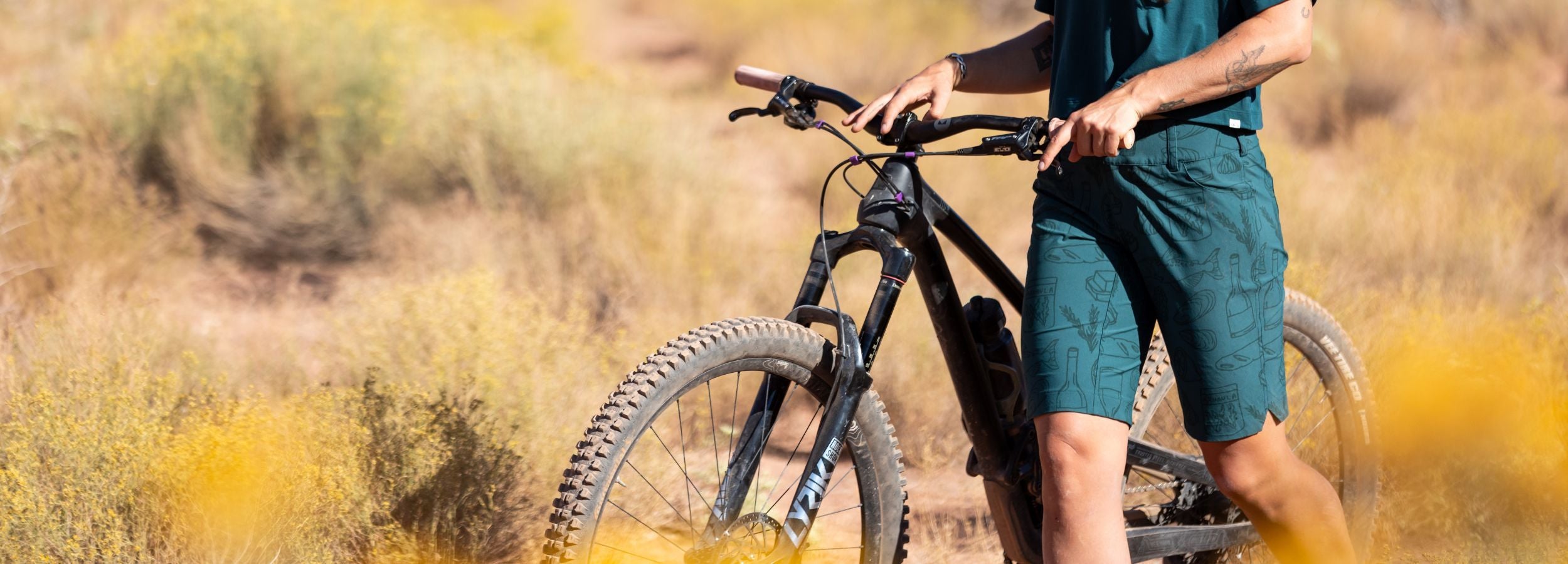 Person standing next to a mountain bike in a desert-like setting with yellowish-brown grass.