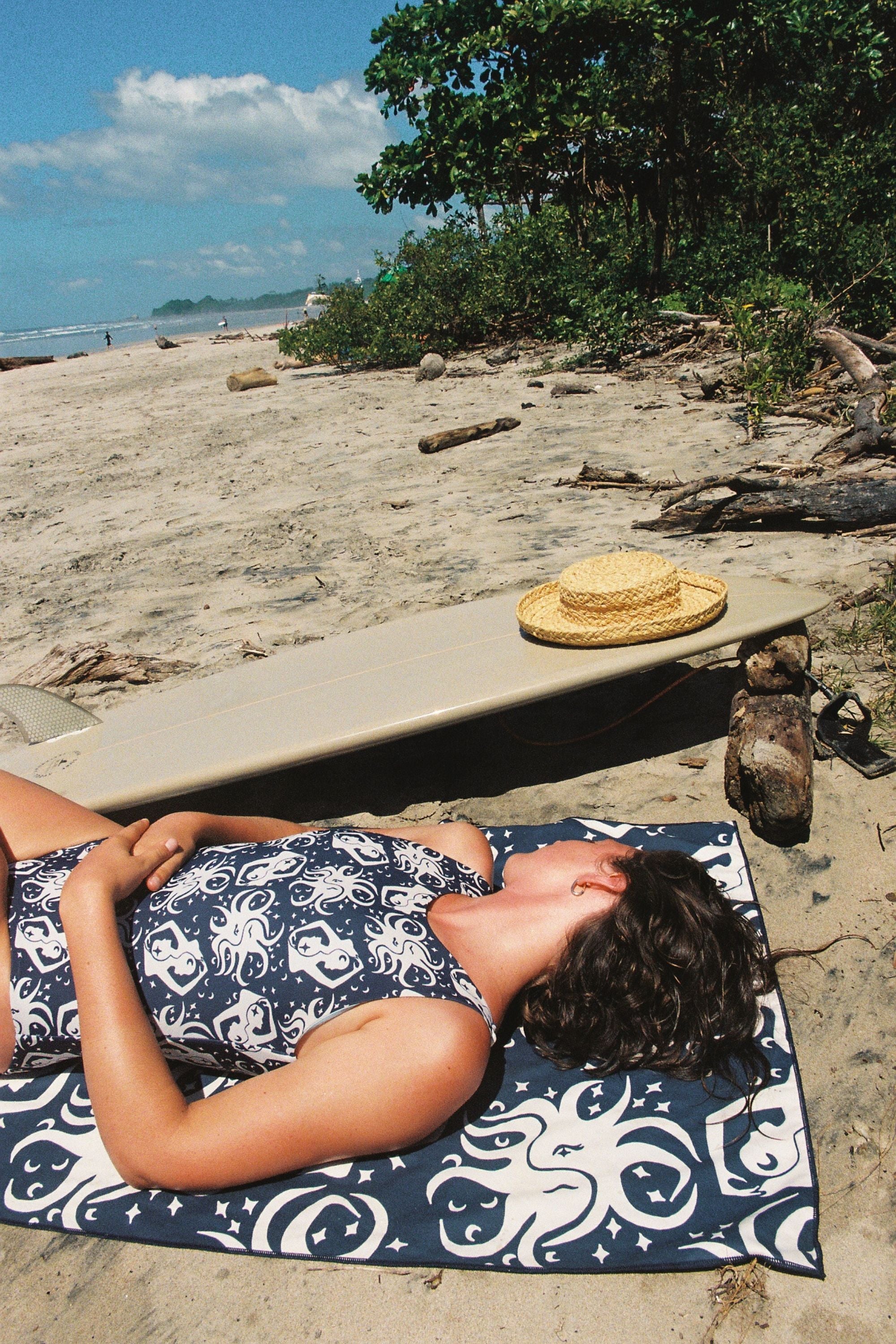 Person lying on a towel at the beach with a hat and surfboard nearby