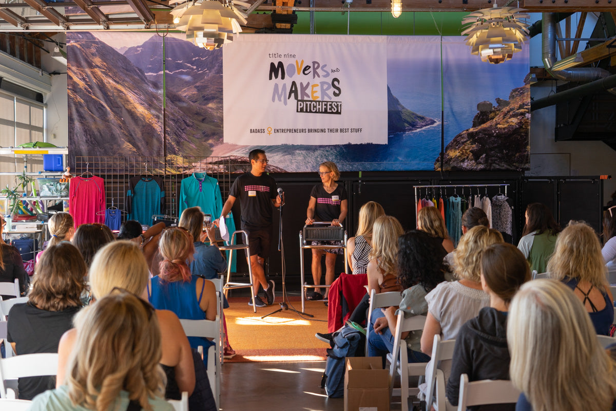 Two presenters at the Title Nine Movers and Makers Pitchfest in front of an audience, with a banner displaying 'Movers and Makers Pitchfest.'