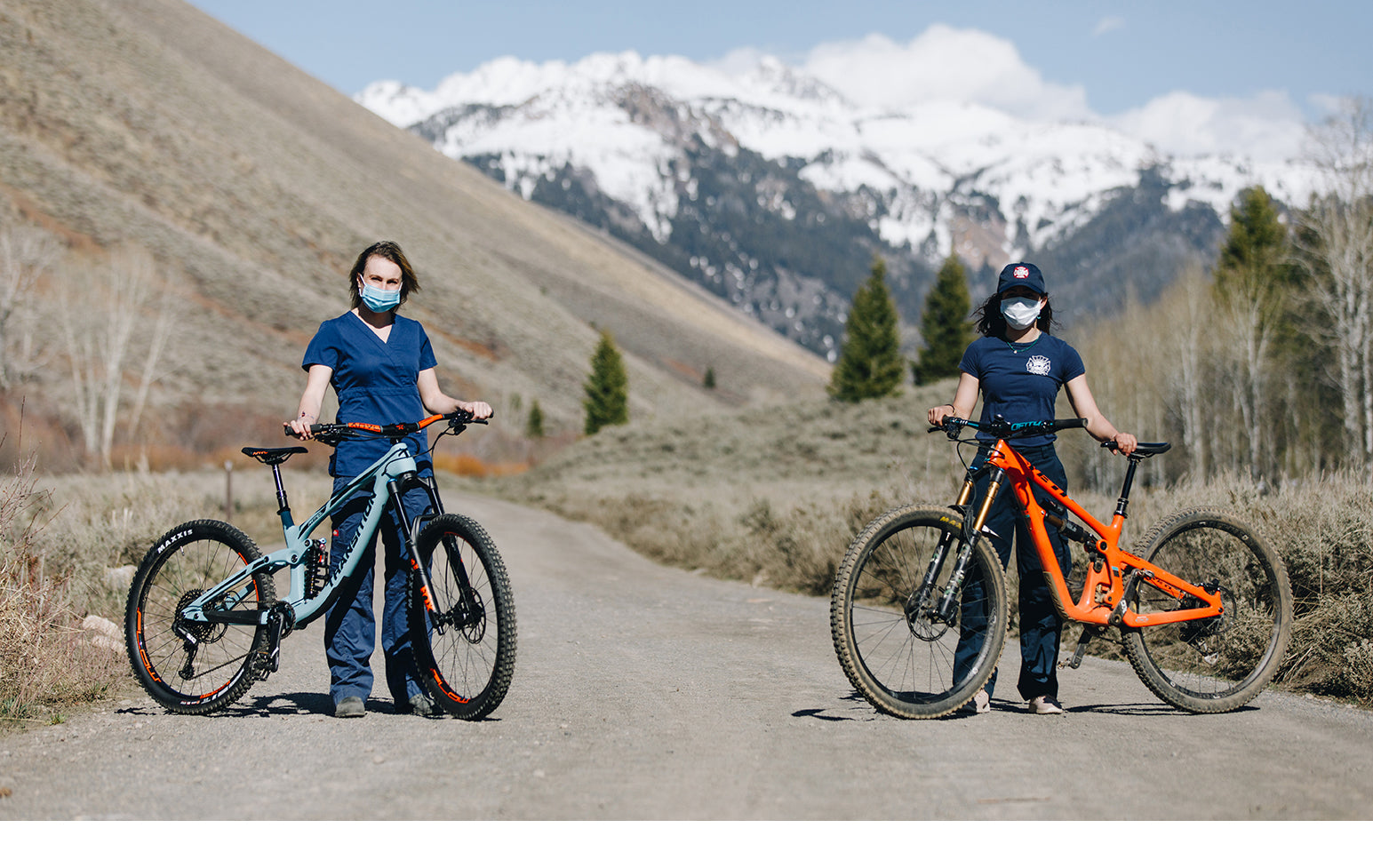 Two women in medical scrubs and masks standing with bicycles on a dirt road with mountains in the background.