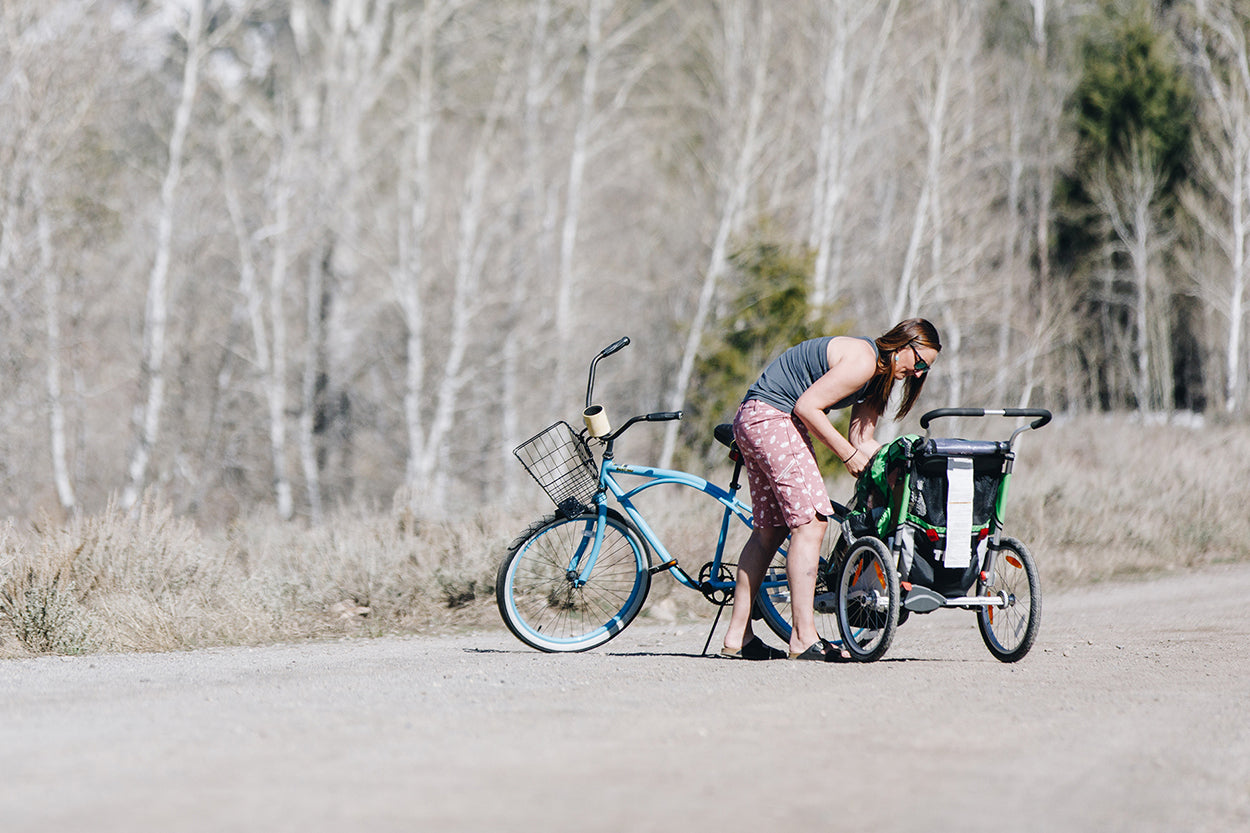 A woman adjusting a child trailer attached to a bicycle on a dirt path.