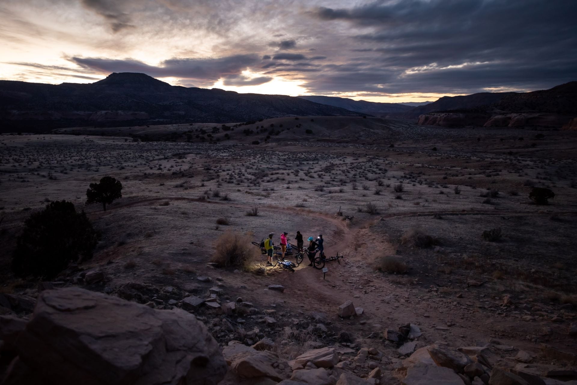 Mountain bikers gathered on a trail at sunset in a desert landscape.