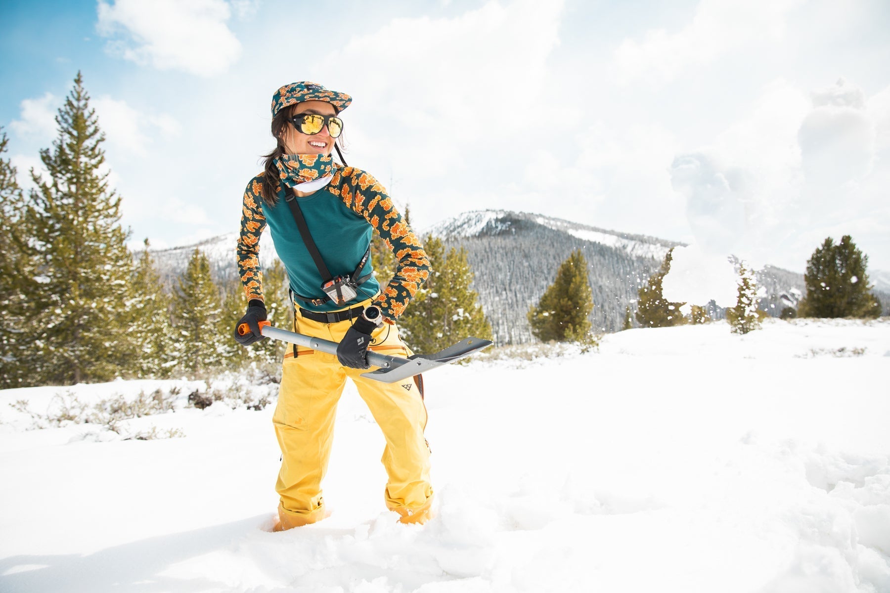 A person in a floral ski outfit standing in snow with a shovel, mountains in the background.