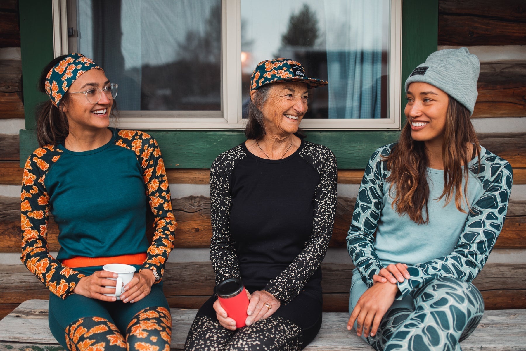 Three women wearing colorful activewear, sitting together outside a cabin, smiling and chatting.