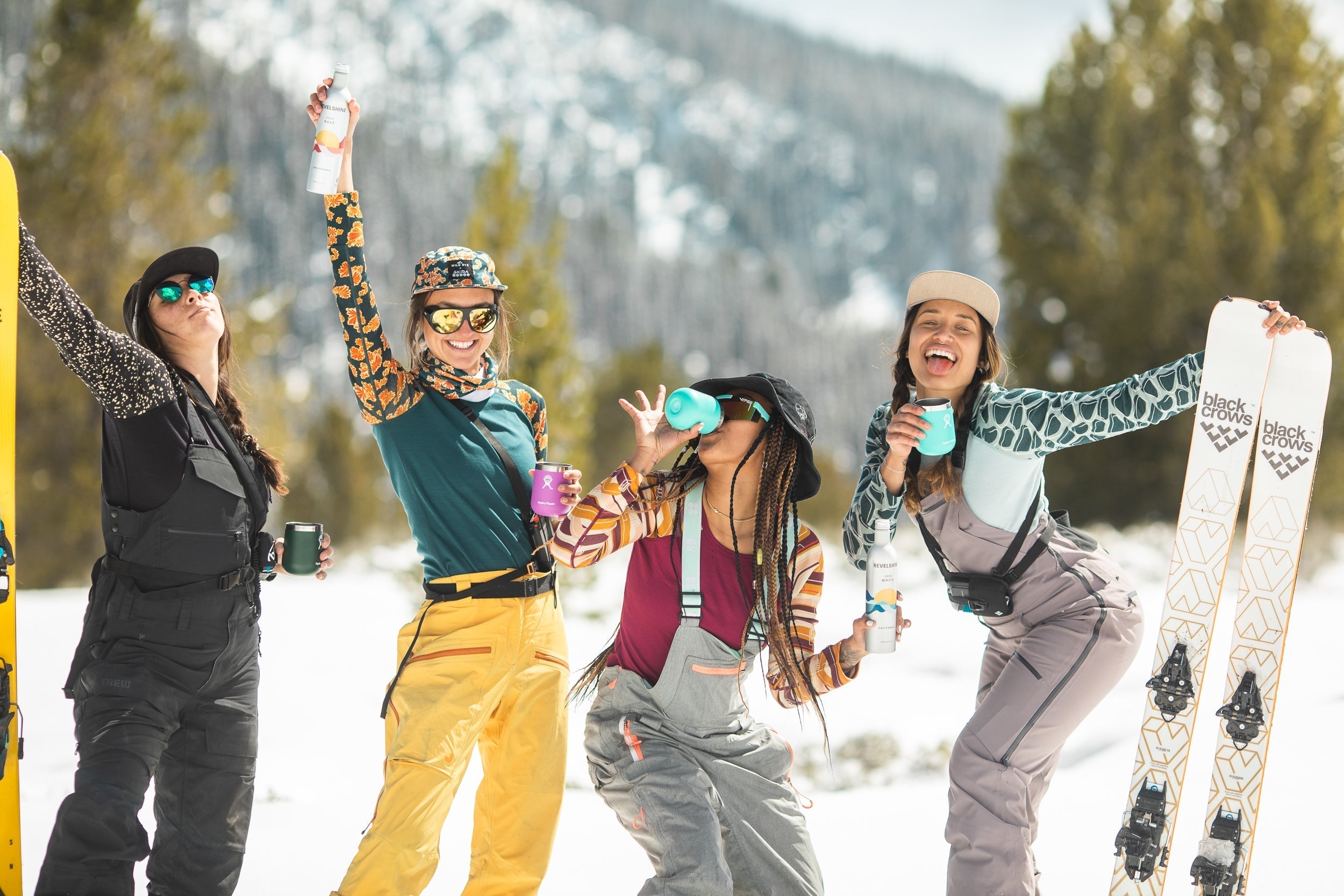 Four women celebrating in winter gear, holding drinks and ski equipment in a snowy landscape.