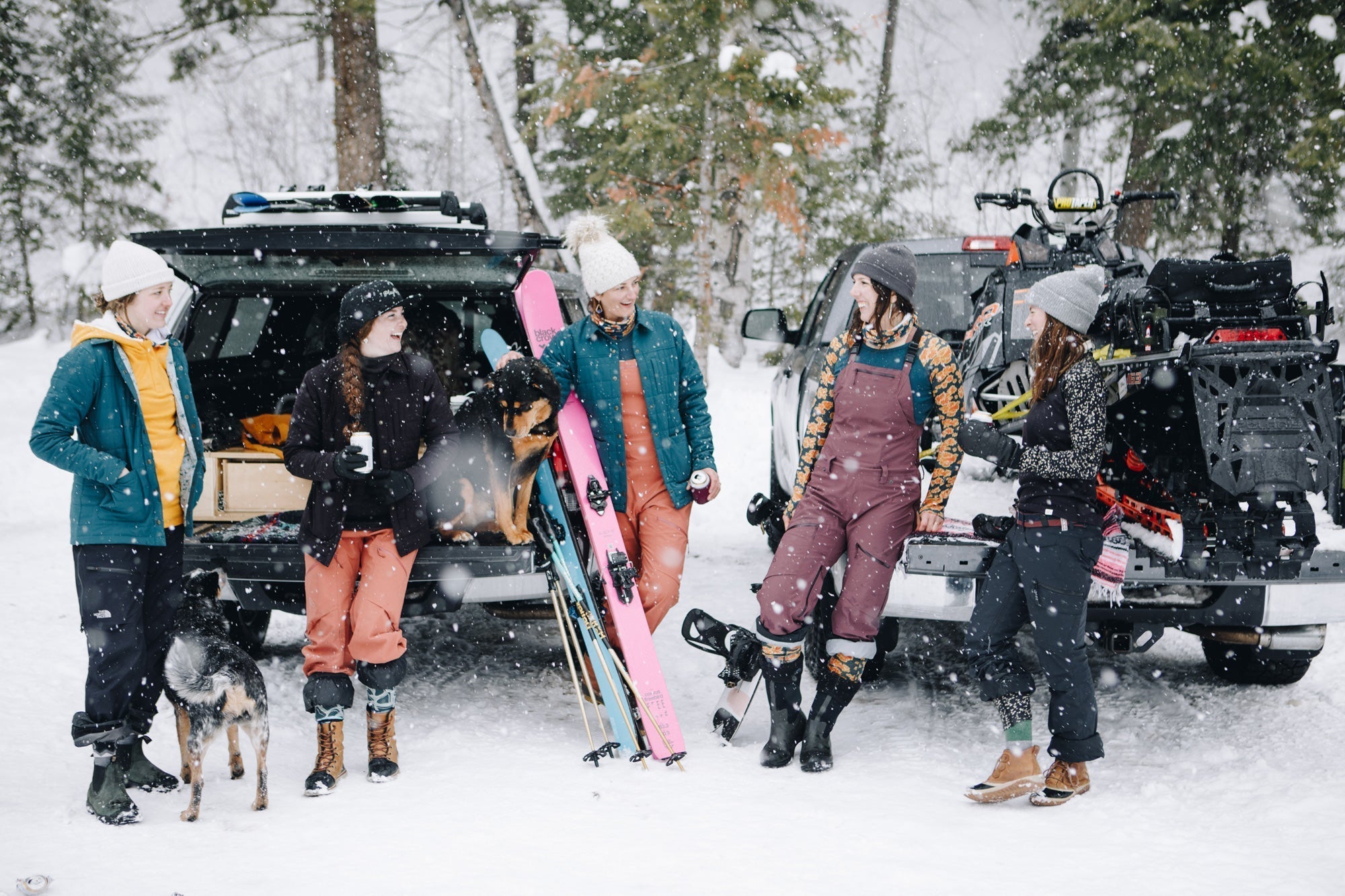 Five women in winter gear standing by a snow-covered vehicle, holding ski equipment and a dog in a snowy landscape.
