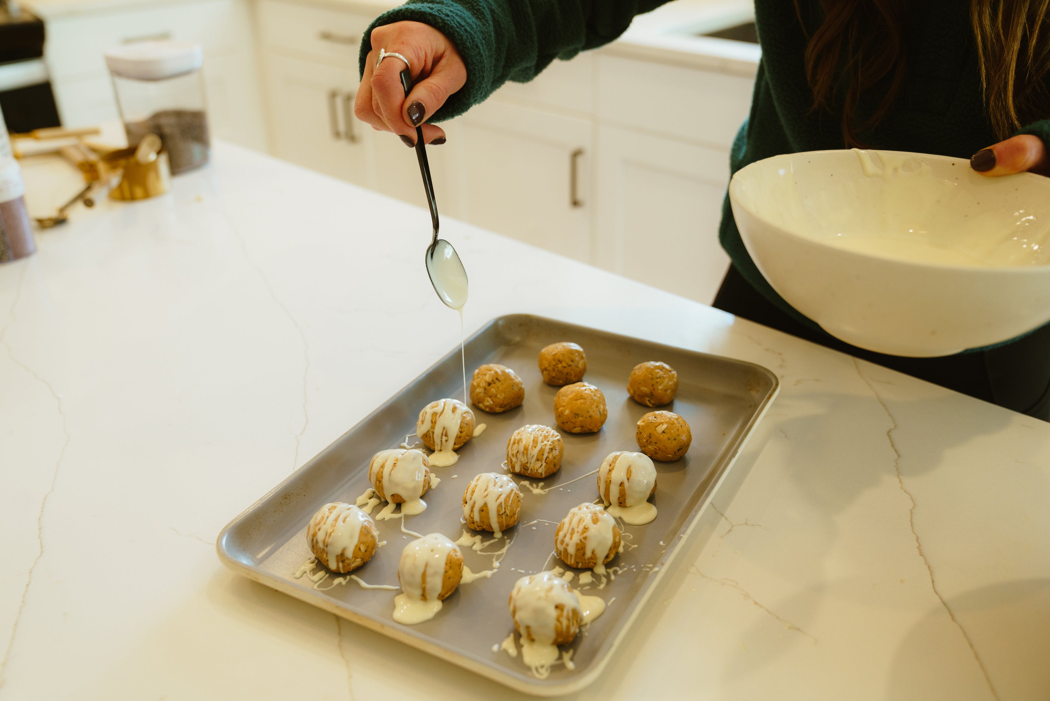 A person drizzling icing over energy balls on a baking tray in a kitchen.