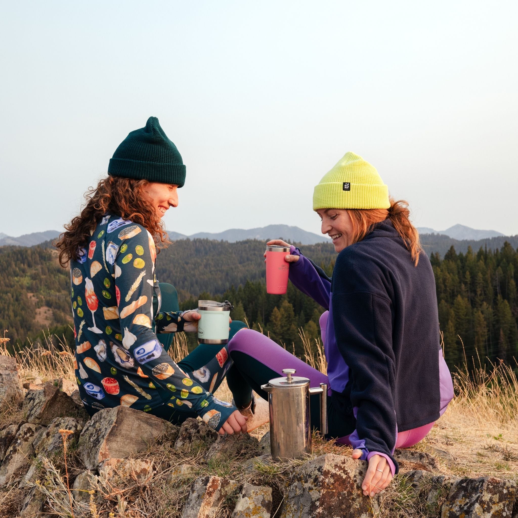 [Spruce Spuntini] Two people sitting on a rock in a mountainous area, enjoying a drink from a french press