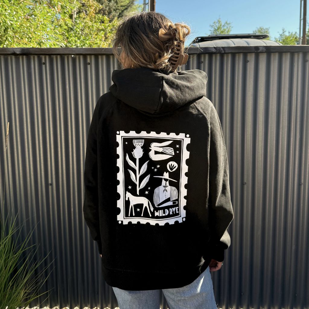 Person wearing a black hoodie with a white graphic design on the back, standing in front of a corrugated metal fence.