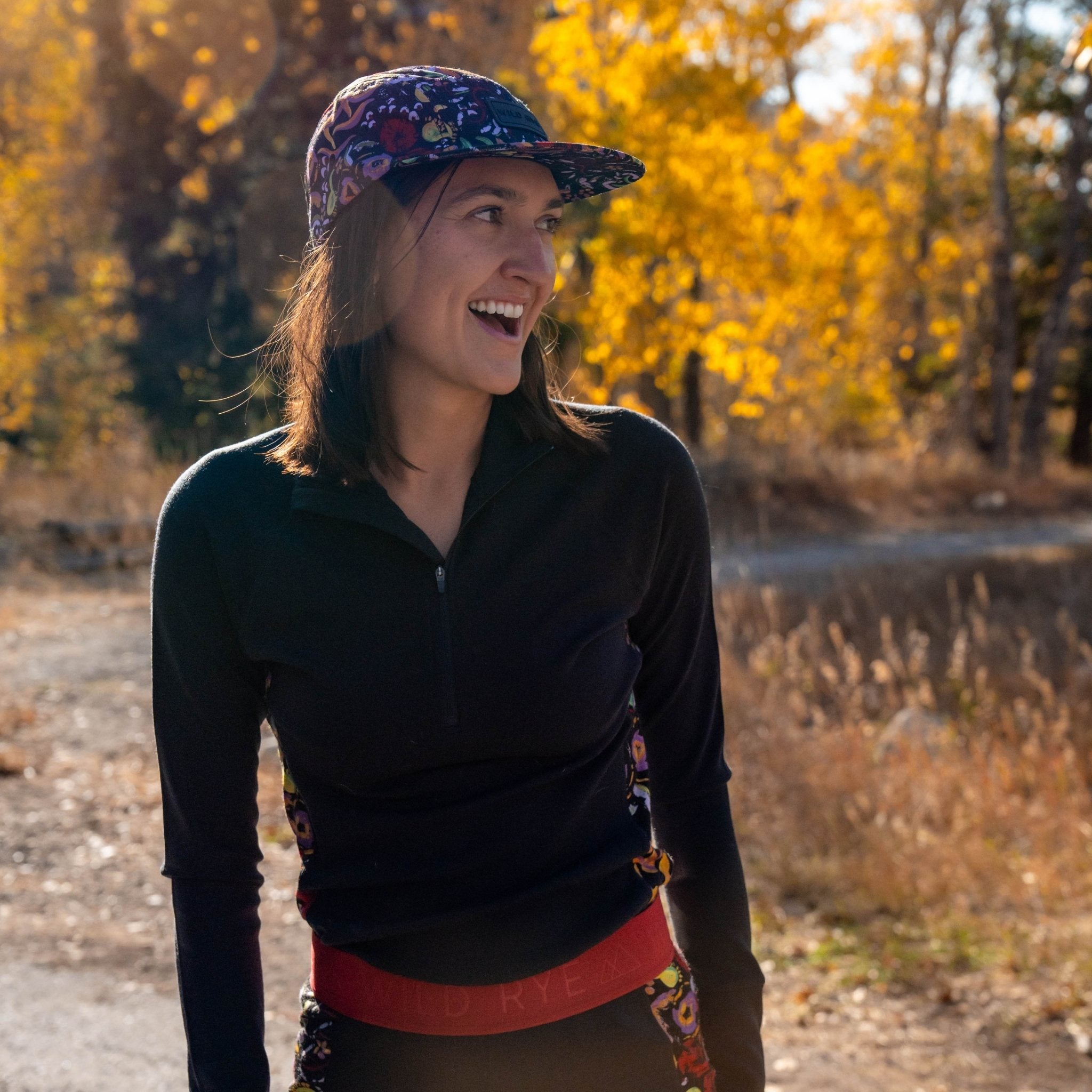 [Understory Blocked] Person wearing a colorful cap and black half zip standing in a natural setting with trees and a body of water.