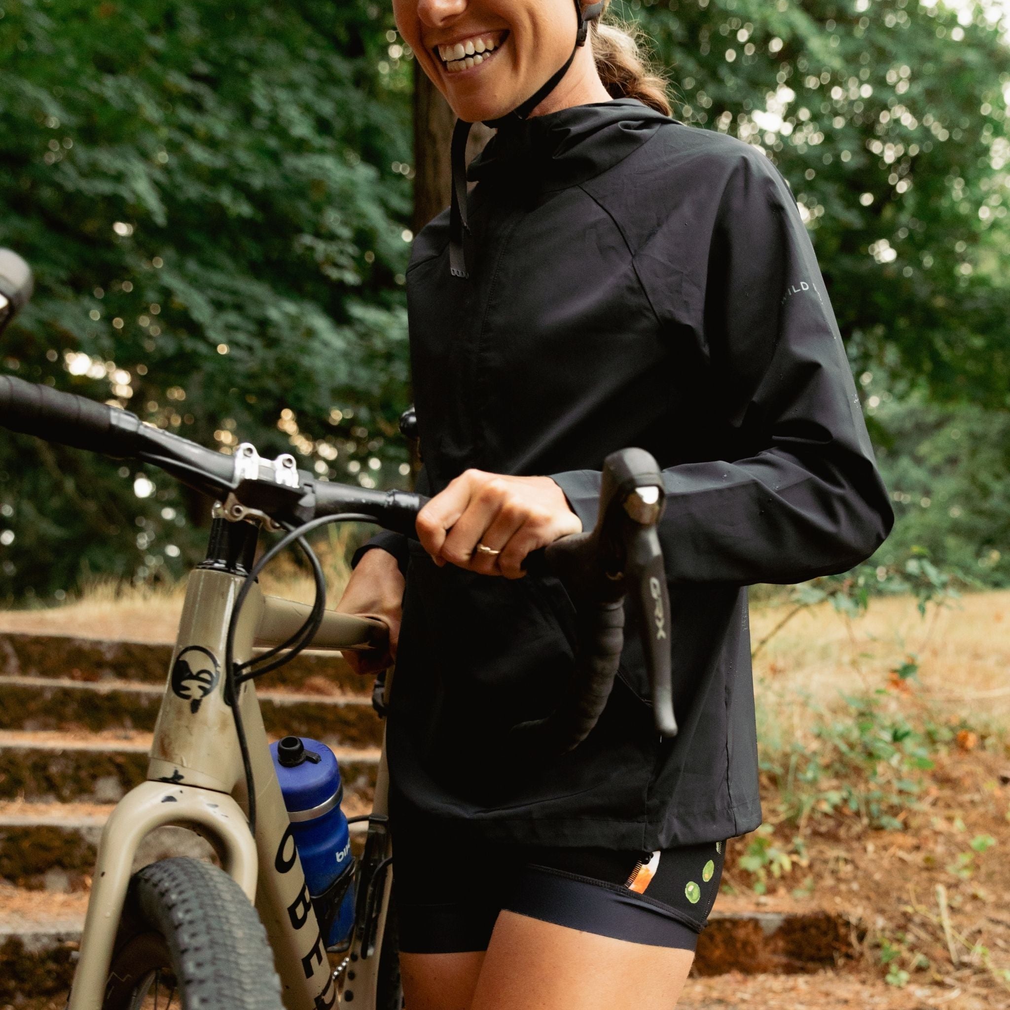 [Black] Person wearing a black jacket and black shorts standing next to a bicycle in a natural setting.