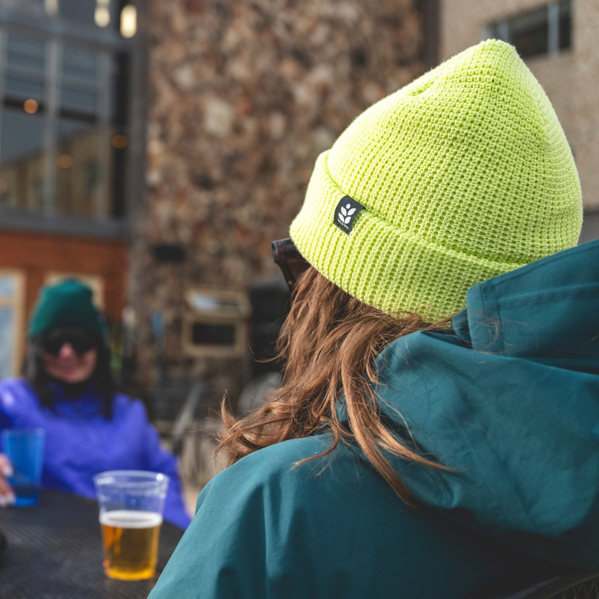[Iced Matcha] Person wearing a bright green beanie and dark green jacket sitting outdoors with another person in the background.