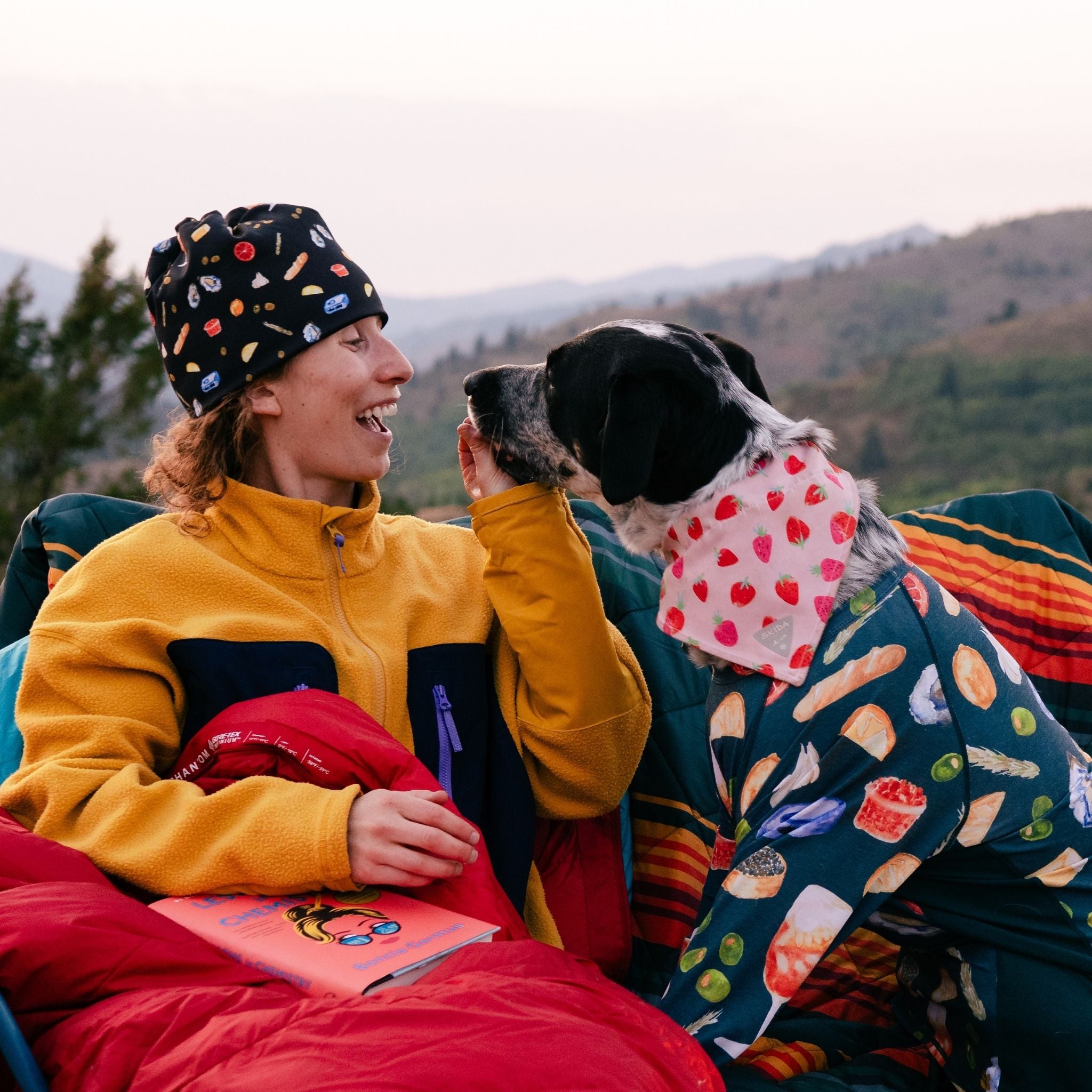 Person snuggled up in a sleeping bag, wearing a yellow jacket and playing with a dog
