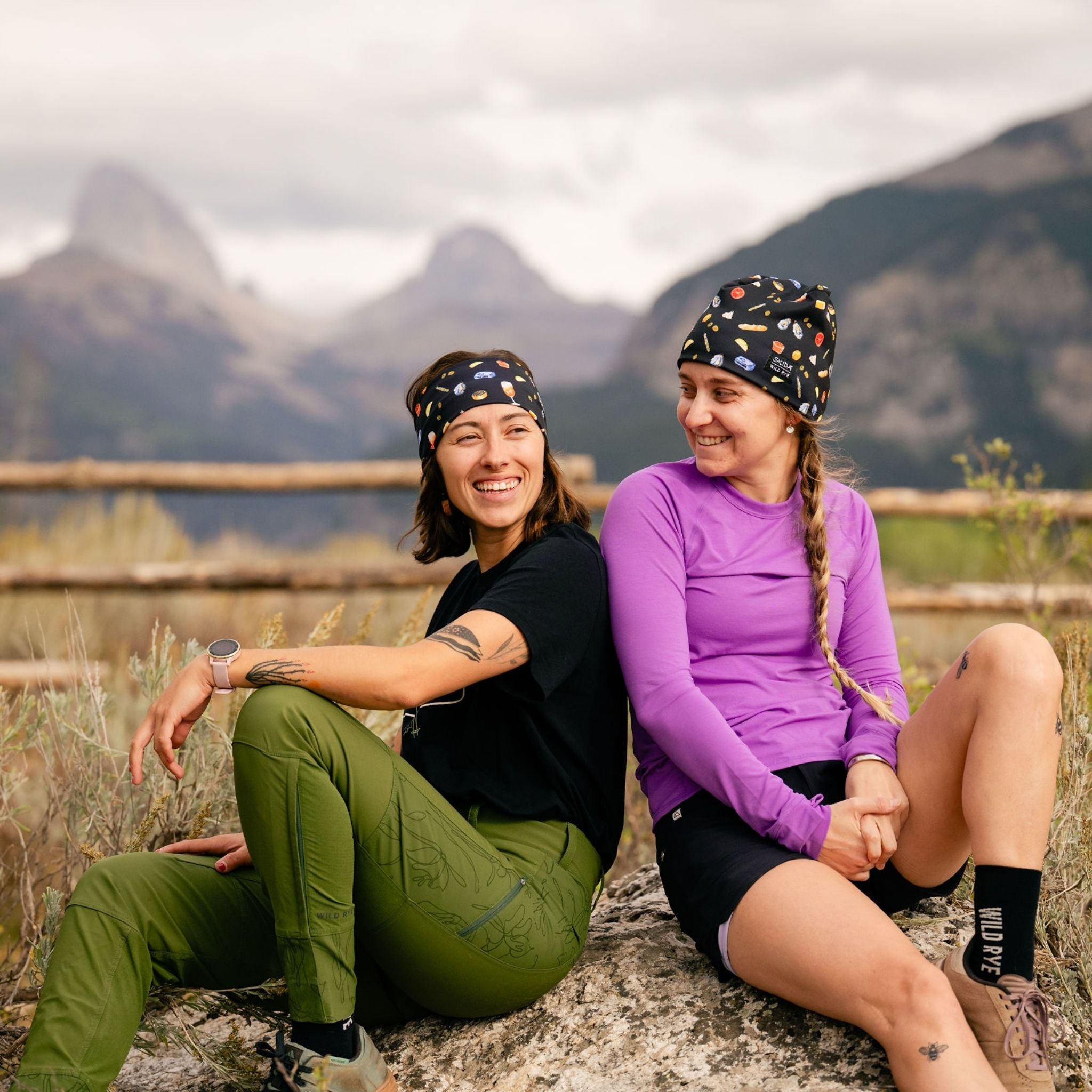 Two women sitting on a rock with mountains in the background, wearing a headband and a hat with food themed print