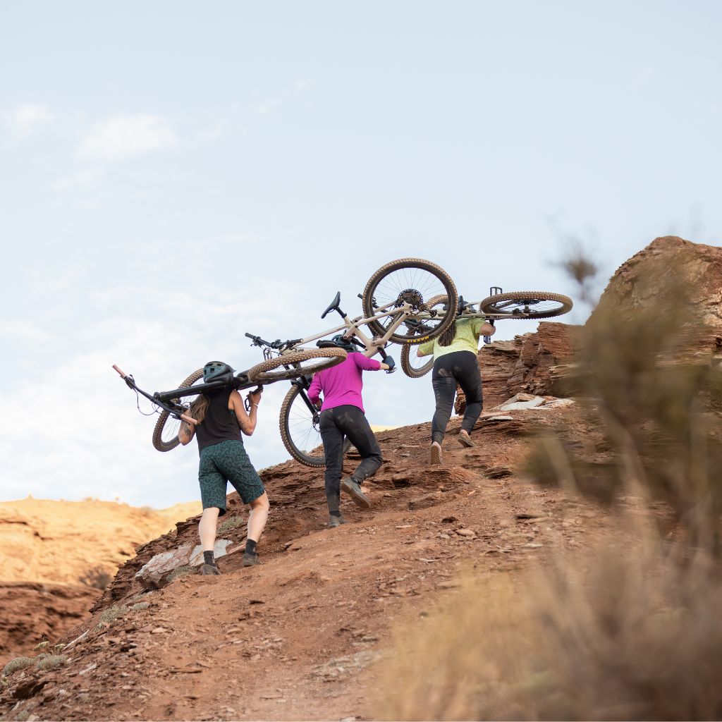 [Spruce Meowww] Women hiking their bikes uphill in the desert wearing Frankie Shorts and Freyah pants