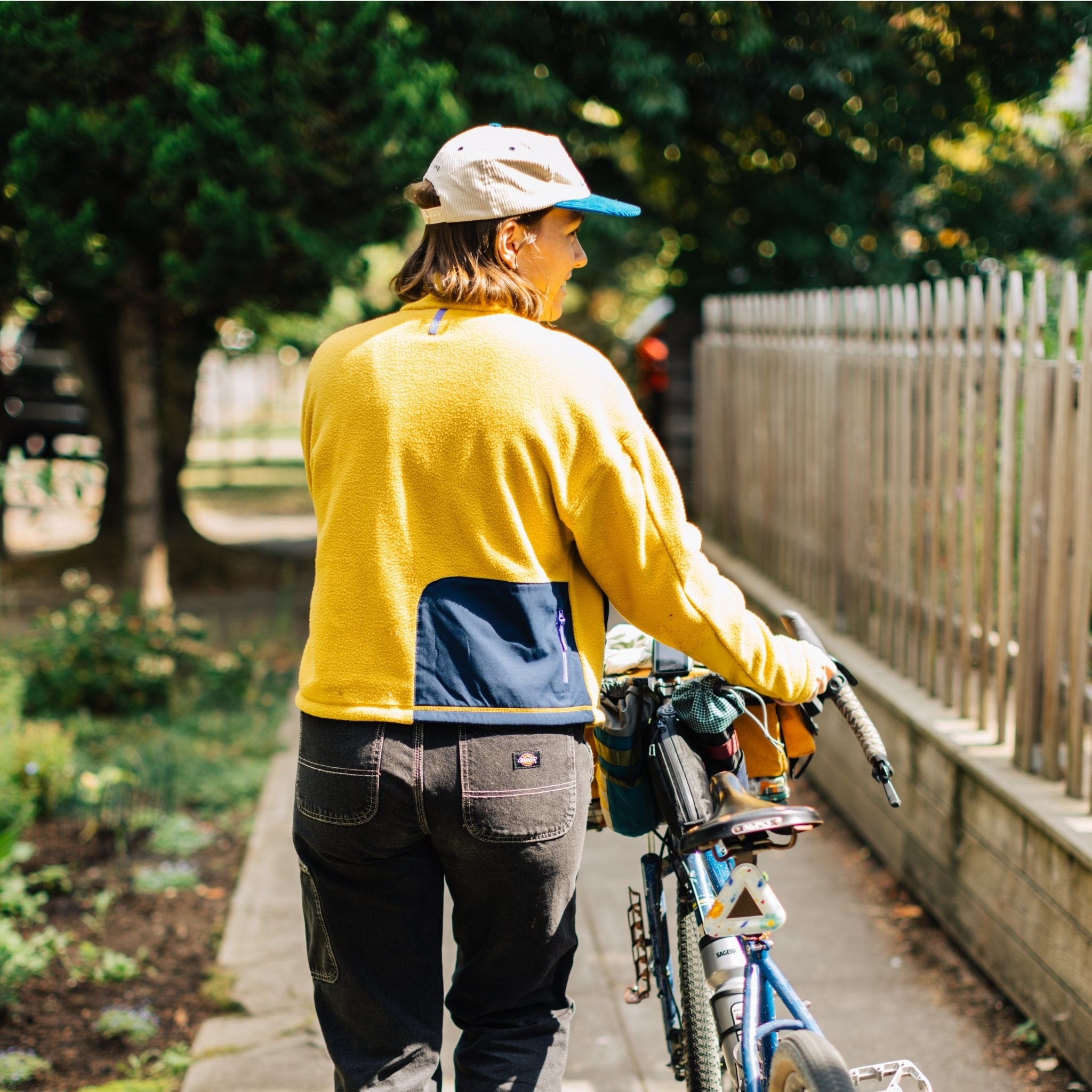 [Saffron] Woman smiling and walking her bike wearing yellow fleece, back view