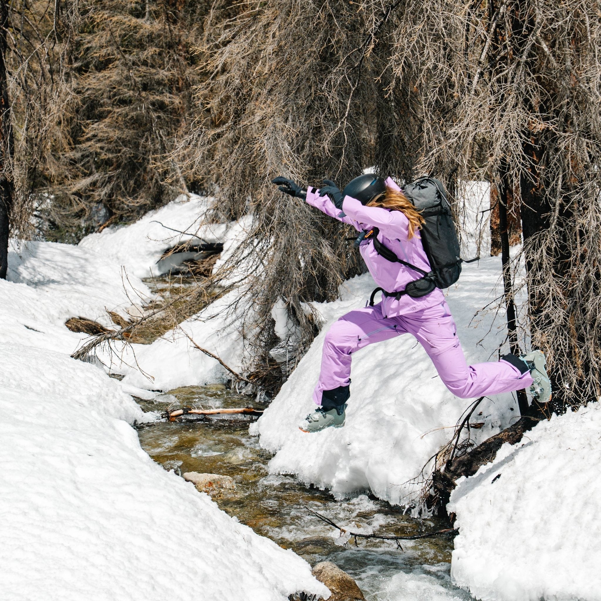 [Amethyst] Person in an amethyst heyburn kit leaping across a running creek in the snowy backcountry