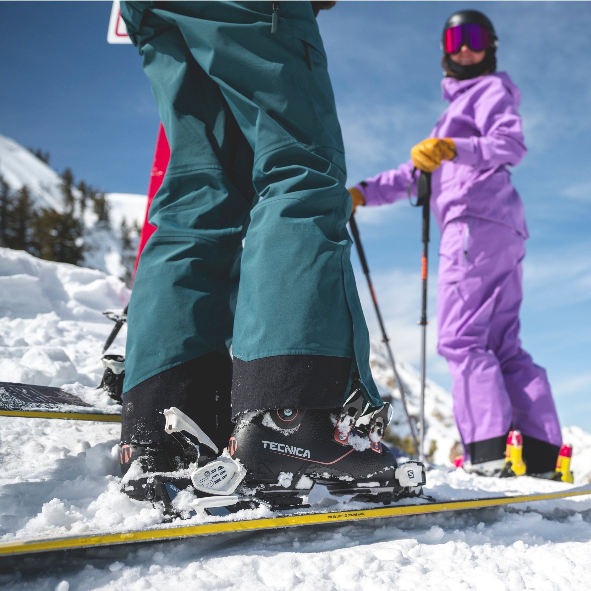 [Shaded Spruce] Two skiers on a snowy mountain with Tecnica ski boots and purple and green outfits.