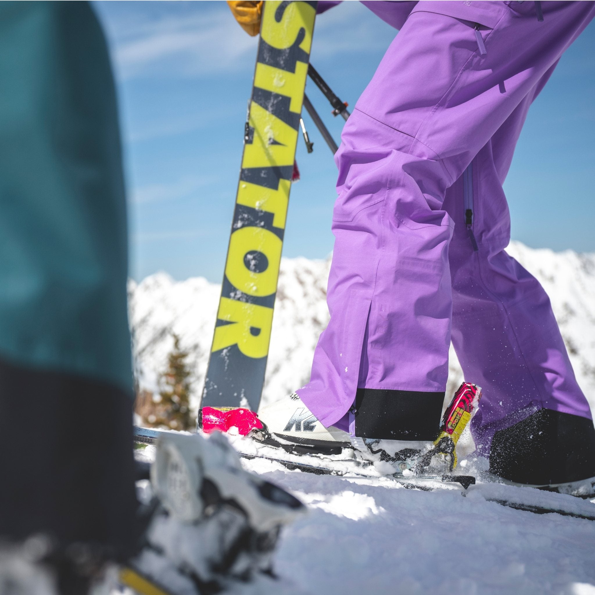 [Amethyst] Person in heyburn bibs in amethyst standing on a snowy slope with skis.