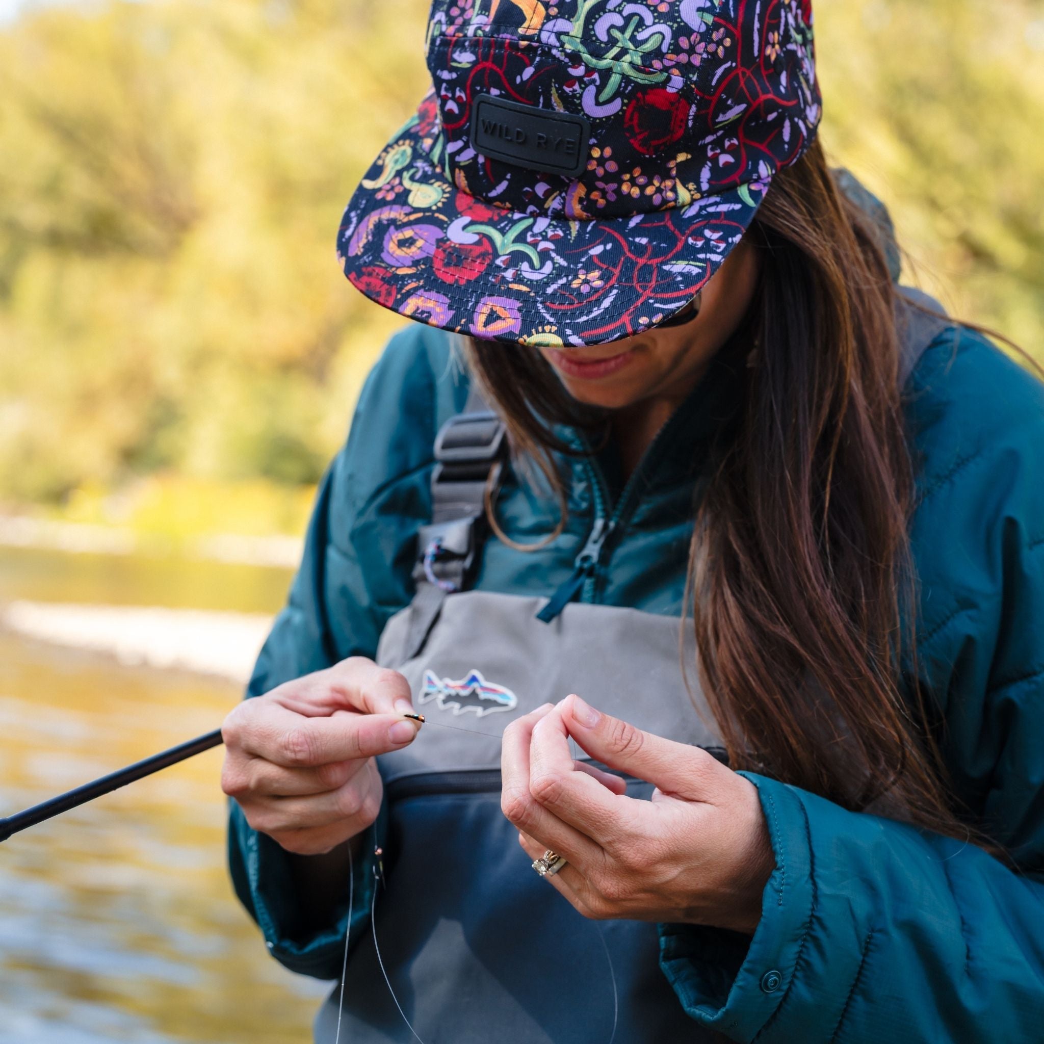 [Shaded Spruce] Person fishing by a body of water wearing a colorful cap and spruce green jacket.