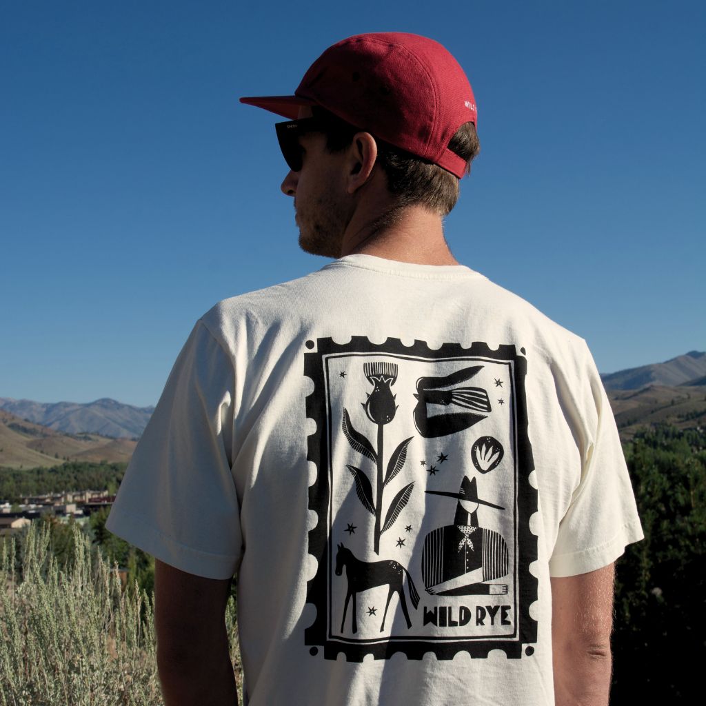 Person wearing a beige t-shirt with a black graphic design and 'WILD RYE' text, standing outdoors with mountains in the background.
