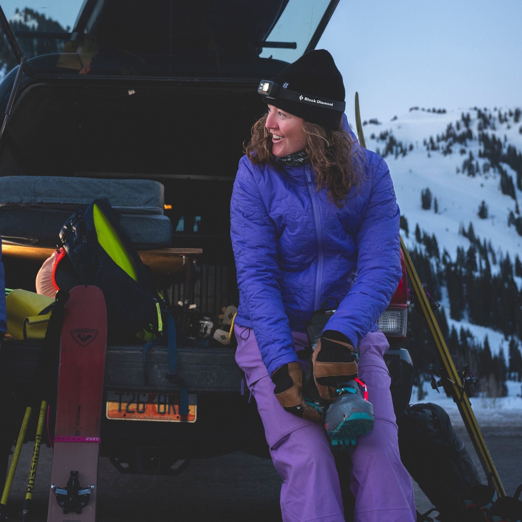 [Iris] Person in winter gear sitting by an open car trunk with skis, in a snowy mountain setting.