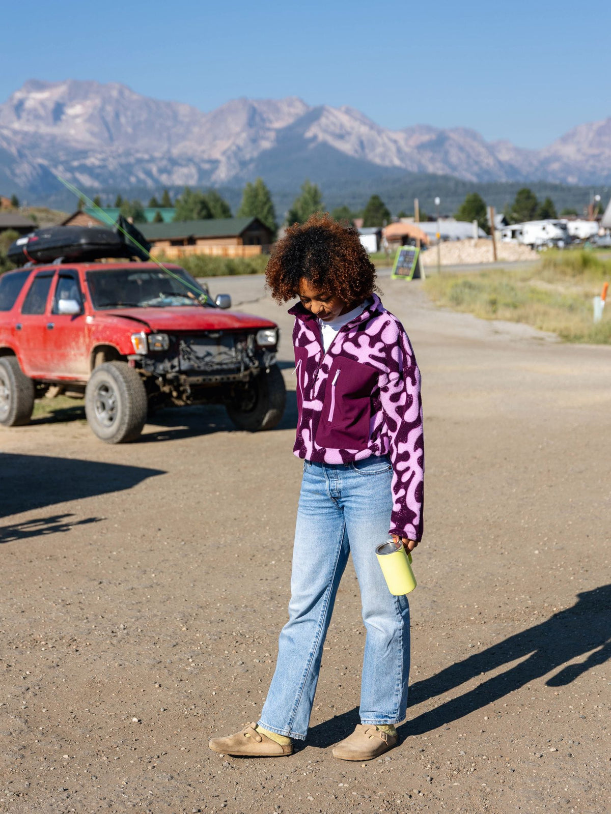 [Mulberry Reef] Lifestyle shot of a model wearing the Banner 3/4 Zip Jacket in purple and pink abstract-print fleece, standing outdoors near mountains