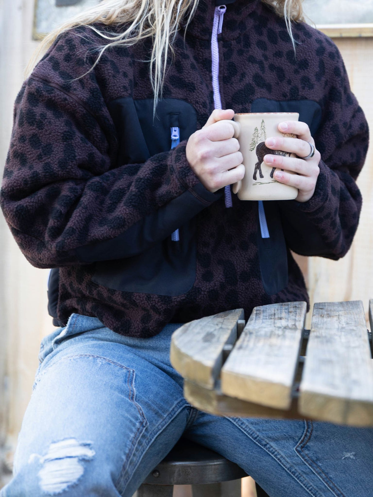 [Dapple] Lifestyle shot of a model wearing the Banner 3/4 Zip Jacket in brown leopard-print fleece, seated outdoors holding a mug
