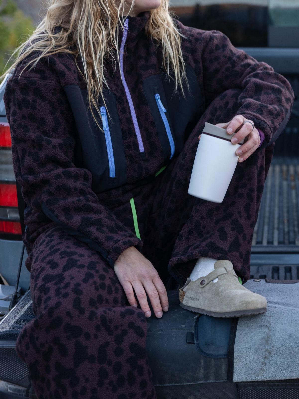 [Dapple] Lifestyle close-up of a model wearing the Banner Pant in brown leopard-print fleece with the matching zip jacket, seated in a truck bed holding a cup